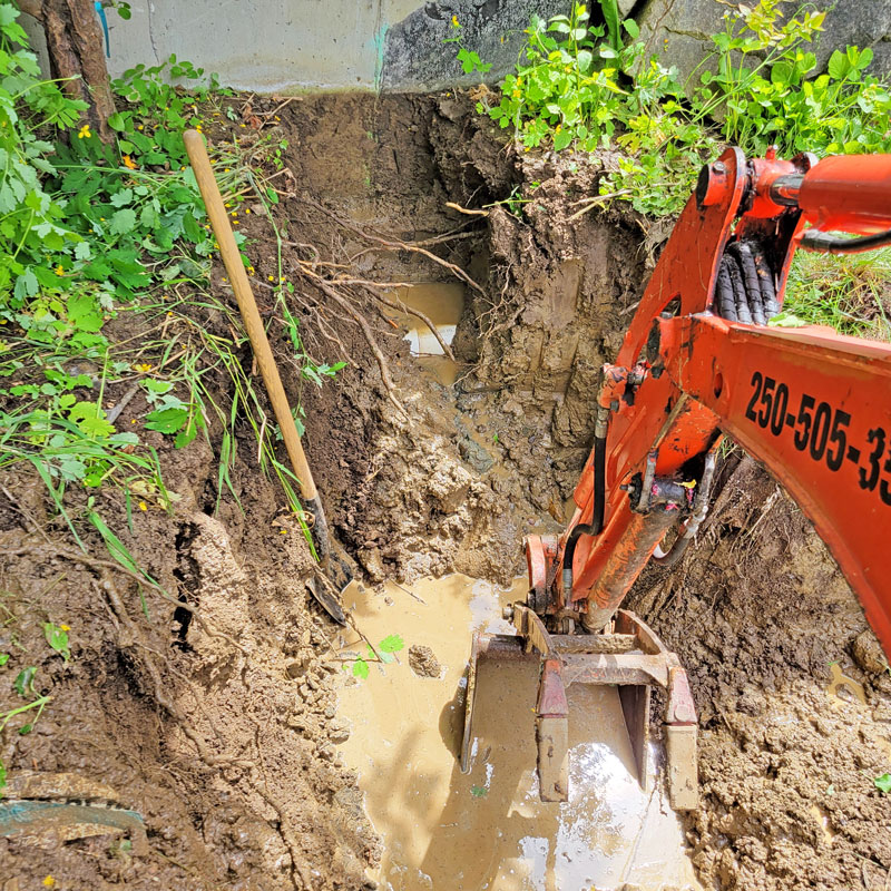 Scoop mini excavator carefully digging along a residential home foundation to install perimeter French drains and waterproofing.<br />